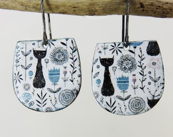 May include: A pair of white enamel earrings with a black cat and floral design. The earrings are shaped like a teardrop and have a silver hook.