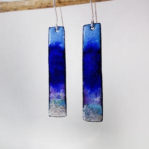 May include: A pair of rectangular earrings with a gradient of blue hues, from light to deep indigo. The earrings are suspended from silver hooks and have a textured, watercolor-like appearance. The bottom edges have a silver finish.