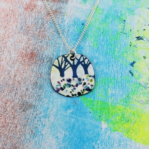 May include: A silver chain necklace with a round pendant featuring a white background with a black silhouette of three trees and a colourful, speckled design.