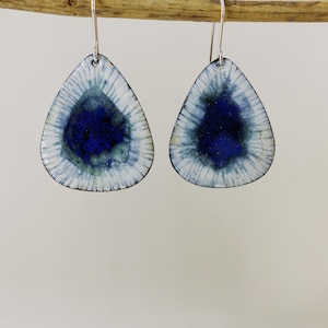 May include: A pair of teardrop-shaped earrings with a white enamel background and a blue enamel centre. The earrings have a silver hook.