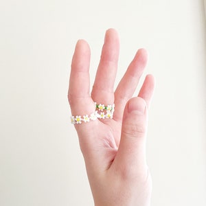 May include: A hand wearing two daisy-shaped beaded rings and a silver chain bracelet.