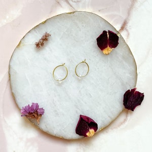 May include: A pair of gold hoop earrings with a small clear stone accent on a white marble surface.