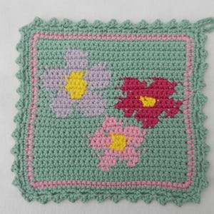 Könnte beinhalten: Ein grüner gehäkelter Topflappen mit einem Blumenmuster in Rosa, Lila und Gelb.