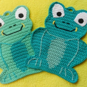 Könnte beinhalten: Zwei gehäkelte grüne Frosch-Topflappen mit weißen Augen und schwarzen Pupillen. Die Frösche haben ein gelbes Lächeln und eine schwarze Nase.