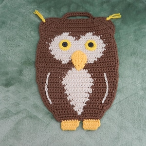 Könnte beinhalten: Ein brauner, weißer und gelber gehäkelter Topflappen in Eulenform mit einer Schlaufe zum Aufhängen. Die Eule hat große gelbe Augen, einen gelben Schnabel und gelbe Füße. Der Topflappen ist aus Garn.