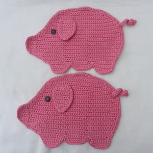Könnte beinhalten: Zwei rosa gehäkelte Schweine-Topflappen mit schwarzen Knopfaugen.