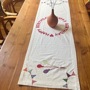 Könnte beinhalten: Ein rechteckiger Tischläufer aus weißem Stoff mit einem kreisförmigen, roten "HAPPY BIRTHDAY"-Schriftzug und einem Herzsymbol. Der Läufer ist mit gestickten, bunten Luftballons und dreieckigen Wimpelketten in verschiedenen Mustern verziert.