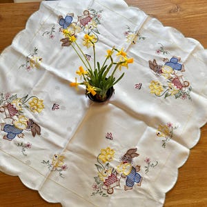 Könnte beinhalten: Cremefarbene, bestickte Tischdecke mit gewelltem Rand. Sie zeigt farbenfrohe, gestickte Bilder von Hasen, Küken und Blumen. Ein kleiner Topf mit gelben Narzissen steht in der Mitte des Tisches und sorgt für einen frühlingshaften Akzent.