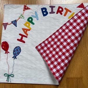 Può includere: Un panno quadrato in tessuto bianco con testo ricamato colorato che recita HAPPY BIRTH, una ghirlanda di piccoli gagliardetti triangolari e due palloncini. L'angolo del panno è ripiegato per rivelare un motivo a quadretti rosso e bianco.