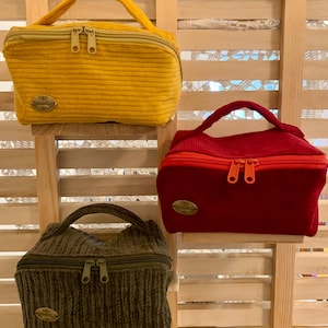 Könnte beinhalten: Drei kleine Kosmetiktaschen aus Cord in Gelb, Rot und Olivgrün. Jede Tasche hat einen oberen Griff, einen Doppelreißverschluss und ein kleines, ovales Emblem. Die Taschen werden auf einem Holzregal ausgestellt.