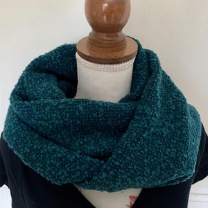 Puede incluir: Bufanda infinita de color verde azulado, hecha de un material suave y texturizado. La bufanda está drapeada alrededor de una cabeza de maniquí, mostrando su diseño circular y su color intenso. La bufanda parece cálida y acogedora.