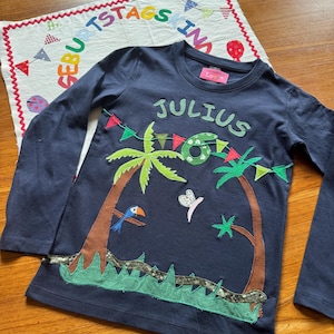 Könnte beinhalten: Ein dunkelblaues Langarmshirt mit einer Applikation aus zwei Palmen, einer grünen Zahl sechs, einem Tukan, einem Schmetterling und einer bunten Wimpelkette. Der Name JULIUS ist in hellgrünen Buchstaben über der tropischen Szene aufgestickt.