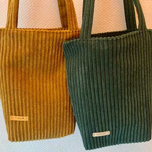 May include: Two tote bags made of corduroy fabric. One bag is mustard yellow and the other is forest green. Both bags have a small leather tag with the word "Everyday" on it.