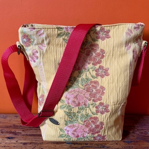 Może przedstawiać: Żółta torba na ramię w kwiaty z czerwonym paskiem. Torba ma powtarzający się wzór różowych kwiatów i zielonych liści. Pasek jest regulowany i wykonany z wytrzymałego, tkanego materiału.