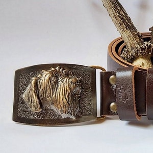 Puede incluir: Un cinturón de cuero marrón con una hebilla de metal que presenta un diseño detallado de cabeza de perro. El cinturón está decorado con dos cuernos.
