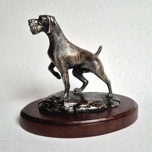 Op de afbeelding: Een bronzen hondensculptuur, mogelijk een jachthondenras, staat op een rotsachtige basis bovenop een ronde, donkere houten basis. De hond staat in een afwachtende houding, met één voorpoot omhoog. De sculptuur heeft gedetailleerde vacht en gelaatstrekken.