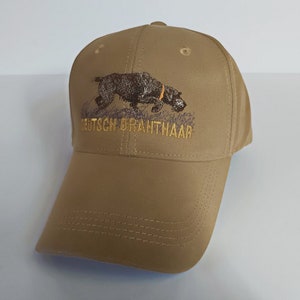 Puede incluir: Una gorra de béisbol de color beige con un perro Braco Alemán de Pelo Duro bordado y el texto "DEUTSCH DRAHTHAAR".