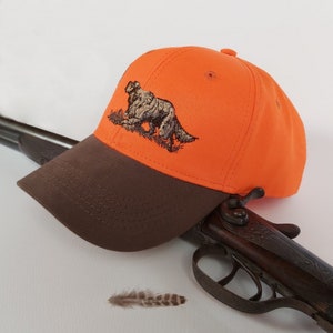 May include: An orange and brown baseball cap with an embroidered image of a hunting dog. The cap is sitting on top of a brown shotgun.