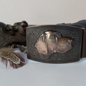 Może przedstawiać: Brązowy skórzany pasek z metalową klamrą, na której znajduje się szczegółowy, tłoczony wzór głowy psa. Głowa psa jest w kolorze miedzi.