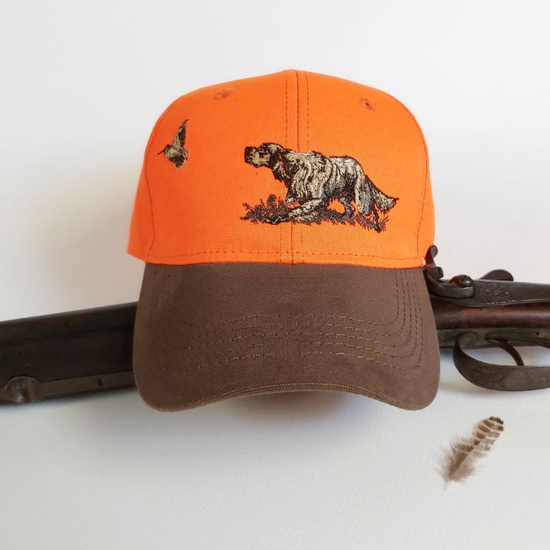 May include: Orange and brown baseball cap with an embroidered image of a hunting dog and a bird. The cap has a brown brim and a bright orange crown.
