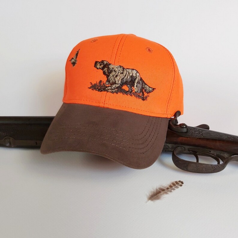 May include: Orange and brown baseball cap with an embroidered image of a hunting dog and a bird. The cap is lying on a white surface next to a shotgun and a feather.
