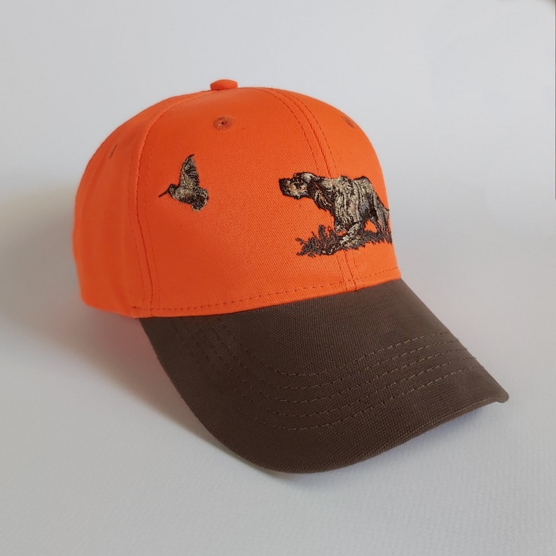 May include: An orange baseball cap with a brown brim. The cap has an embroidered design of a dog and a bird.