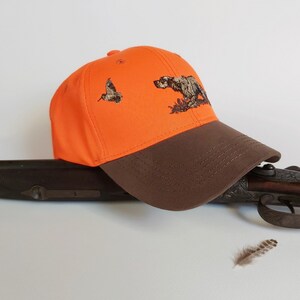 May include: Orange and brown baseball cap with an embroidered hunting dog and bird design.