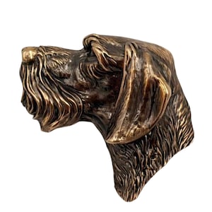 Op de afbeelding: Een bronskleurig metalen sculptuur van een hondenkop in profiel, met een textuur van vacht met zichtbare details, een prominente snuit en een lang, hangend oor. De metallic afwerking vertoont verschillende tinten bruin en goudkleurige accenten.