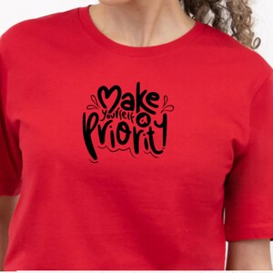 K&ouml;nnte beinhalten: Rotes T-Shirt mit einer schwarzen Grafik, auf der "Mach dich selbst zur Priorit&auml;t" mit einem Herzsymbol steht.