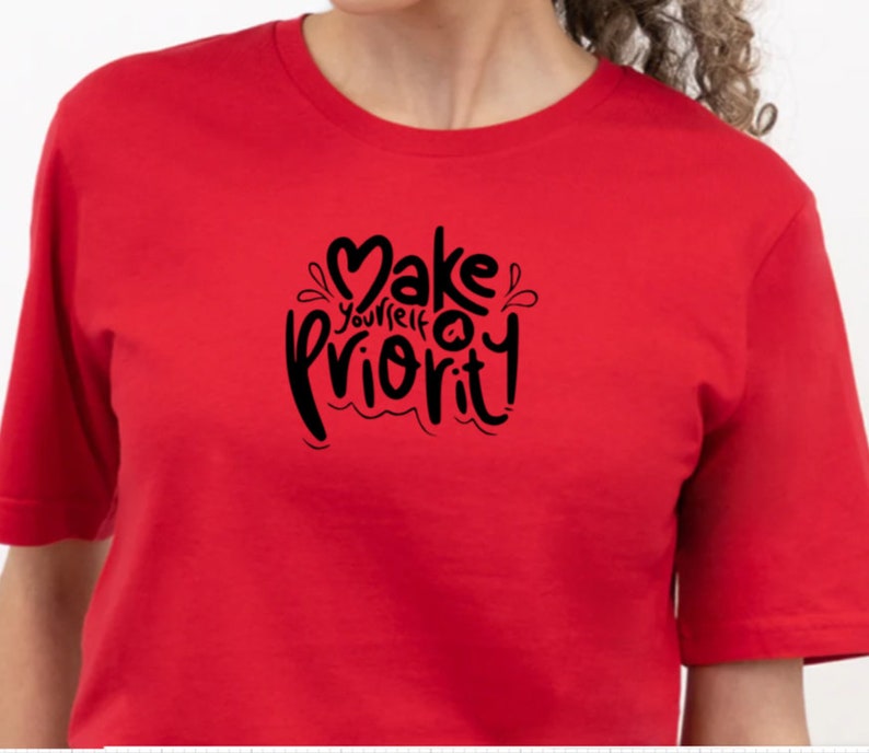 K&ouml;nnte beinhalten: Rotes T-Shirt mit einer schwarzen Grafik, auf der "Mach dich selbst zur Priorit&auml;t" mit einem Herzsymbol steht.