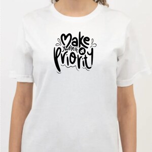 K&ouml;nnte beinhalten: Wei&szlig;es T-Shirt mit schwarzem Text, der "Mach dich zur Priorit&auml;t!" lautet.