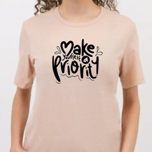 K&ouml;nnte beinhalten: Ein hellrosa T-Shirt mit einer schwarzen Grafik, auf der in einer stilisierten Schrift "Mach dich selbst zur Priorit&auml;t" steht.