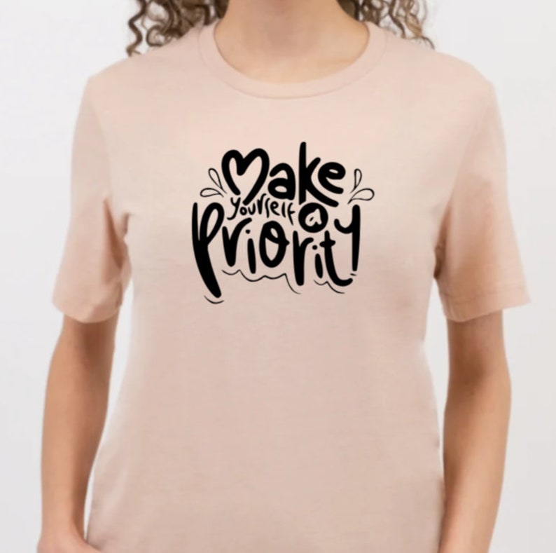 K&ouml;nnte beinhalten: Ein hellrosa T-Shirt mit einer schwarzen Grafik, auf der in einer stilisierten Schrift "Mach dich selbst zur Priorit&auml;t" steht.