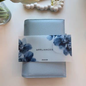 May include: A light blue textured leather planner with a rectangular card placed on top. The card features a white background with dark blue floral illustrations on the sides, the text APPLIANCES in the center, and whimsy & wallet at the bottom.