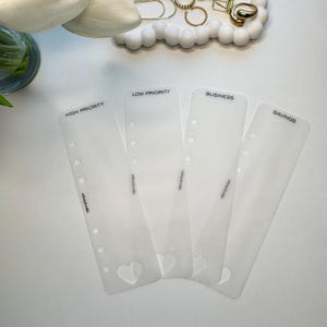 May include: Four translucent plastic planner dividers with punched holes along the left side. Each divider features a printed label at the top reading HIGH PRIORITY, LOW PRIORITY, BUSINESS, and SAVINGS, with a small heart cutout at the bottom.