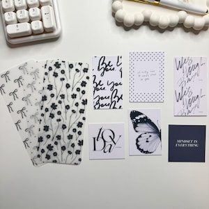May include: A collection of decorative cards and stationery items. Includes cards with floral and bow patterns, inspirational quotes, and a butterfly illustration. The color palette is primarily black and white, with a minimalist aesthetic.
