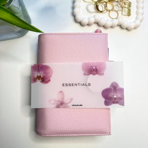 May include: A light pink textured leather planner with a horizontal paper band featuring purple orchid illustrations and the text ESSENTIALS and whimsy & wallet. The planner is positioned vertically with a clean, structured design and a matte finish.