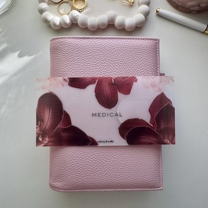 May include: A light pink leather wallet with a textured surface. A clear card with the word "MEDICAL" and a floral design is placed on top. The brand name "whimsy & wallet" is printed on the card.