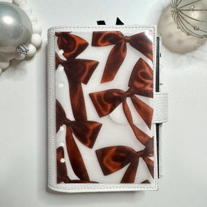 May include: White leather planner with a clear overlay featuring a pattern of brown satin bows. The planner has a snap closure and a ring binder mechanism. Decorative holiday ornaments are in the background.