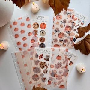 May include: A collection of translucent planner sticker sheets featuring autumn-themed illustrations including pumpkins, pinecones, cookies, mugs, and fall foliage. The sheets are labeled with text such as Pumpkin Season, A Taste of Fall, and All Things Fall.
