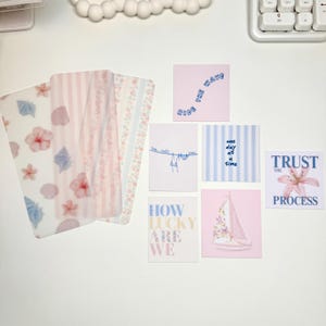 May include: A collection of decorative cards and stationery items. The cards feature various designs, including floral patterns, seashells, and inspirational quotes like "Ride the Wave" and "Trust the Process." The color palette is soft, with pinks, blues, and whites.