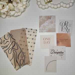 May include: An assortment of decorative cards and bookmarks. The bookmarks feature patterns of bows, dots, and abstract lines. The square cards display inspirational quotes and cloud imagery. Colors include beige, white, and black.