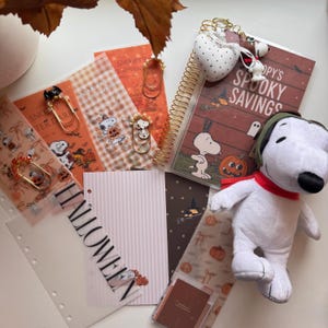 May include: A collection of Halloween-themed stationery including a spiral-bound notebook titled Snoopy's Spooky Savings, a plush Snoopy dog, decorative paper clips with beads, translucent patterned sheets, and a page divider labeled HALLOWEEN.