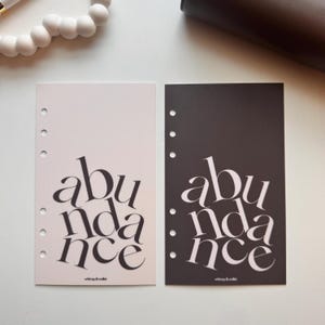 May include: Two rectangular planner inserts with six punched holes along the left side. One insert is light beige with black text, and the other is black with white text. Both feature the word abundance in a large, stylized serif font.