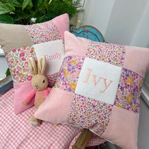 Peut inclure: Deux coussins carrés en patchwork avec des motifs floraux et les noms "Emily" et "Ivy" brodés en rose. Un lapin en peluche rose et beige est placé entre les coussins, pour une touche enfantine.