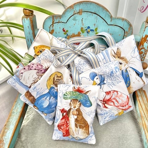 Könnte beinhalten: Ein Set aus acht kleinen Stoffbeuteln mit verschiedenen Peter Rabbit-Charakteren darauf gedruckt. Die Beutel sind aus weißem Stoff mit blau-weißen Streifen und haben ein blau-weiß gestreiftes Band daran befestigt. Die Beutel sind auf einem blau-weiß lackierten Holzstuhl gestapelt.