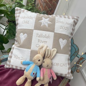 Può includere: Un cuscino patchwork beige e bianco personalizzato con dettagli ricamati, tra cui il testo "Wednesday's Child", "Tallulah River" e "25th June 2025". Due peluche a forma di coniglietto, con giacche blu e rosa, sono appoggiati su una superficie bordeaux.
