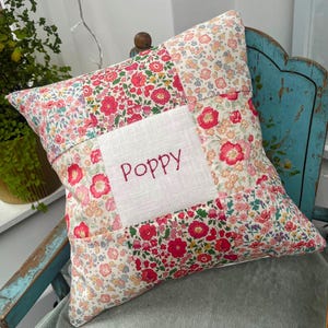 May include: A white cushion with a patchwork design of pink and red floral fabric. The cushion has a white square in the centre with the name "Poppy" embroidered in red.