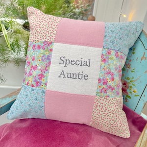 May include: A square patchwork cushion with floral and pink fabric panels. The centre panel has the embroidered text "Special Auntie" in grey. The cushion is placed on a pink velvet surface.