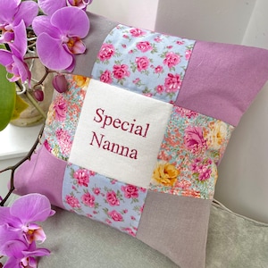 May include: Square patchwork pillow with the embroidered text "Special Nanna". The pillow features a mix of floral and solid fabric squares in shades of pink, lavender, and gray. A sprig of purple orchids is in the background.